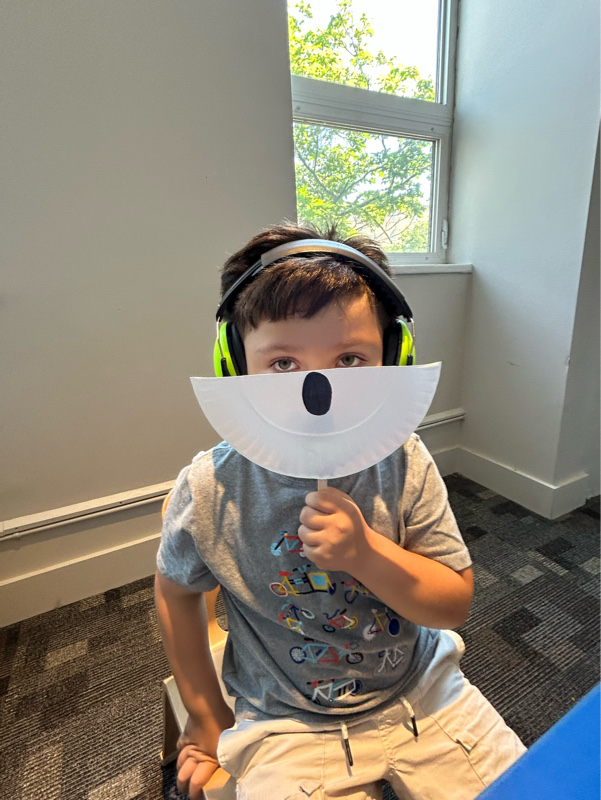 A child wearing headphones, holding a paper mask/visual aid — likely a structured ABA activity (sensory, communication, or play-based learning).
